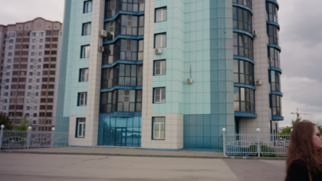 Black gentleman raises shoulder while walking beside elegant companion who strides like model along modern city promenade, moving camera frames tall residential towers and cobblestone pavement