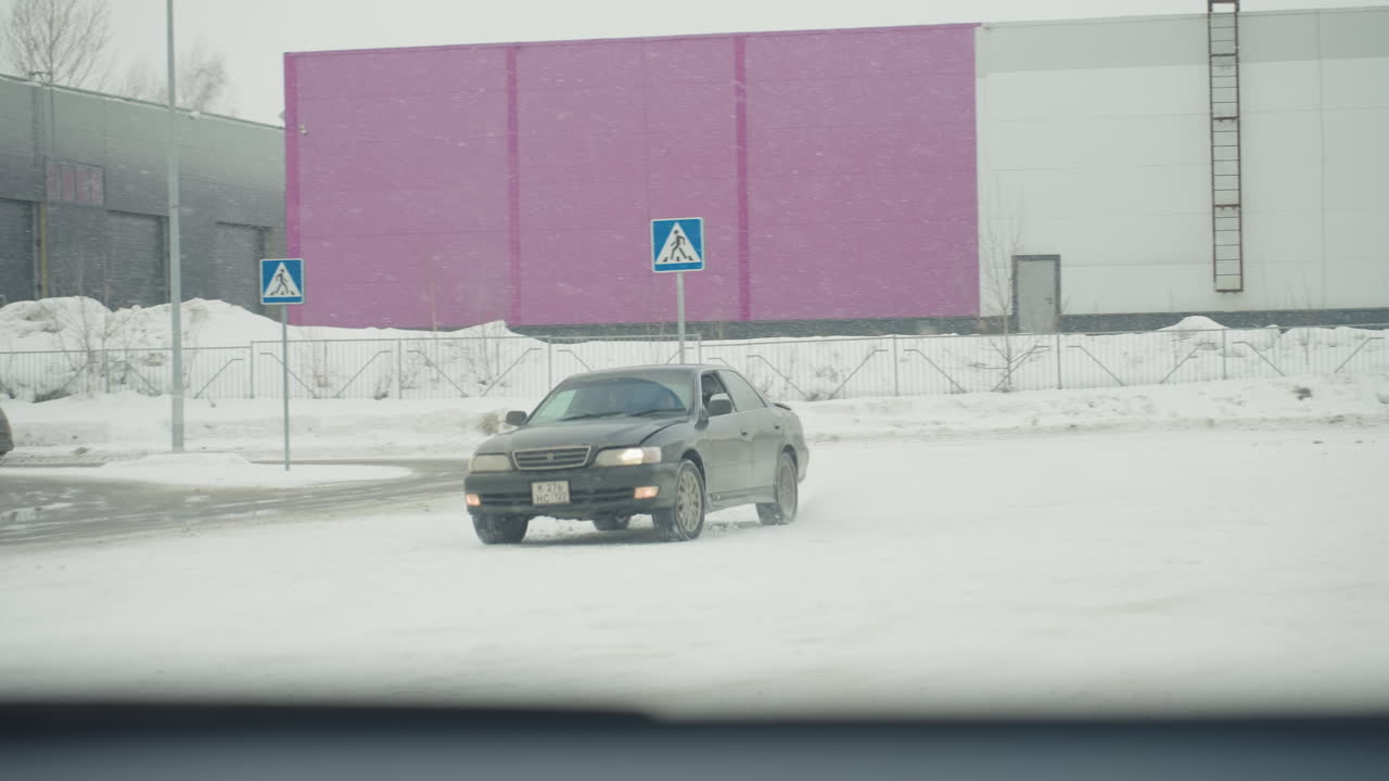 car drives through snowy parking lot past fence and building under falling snowflakes with blurred background showing sign post and industrial structure through window pane