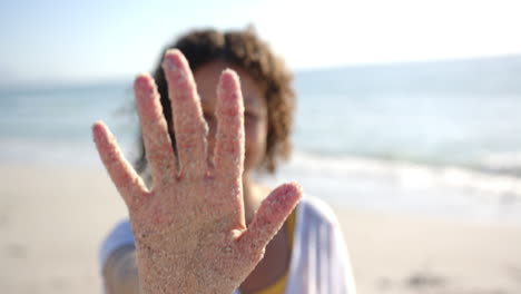 a sandy hand is raised towards the camera, obscuring a person's face at the beach