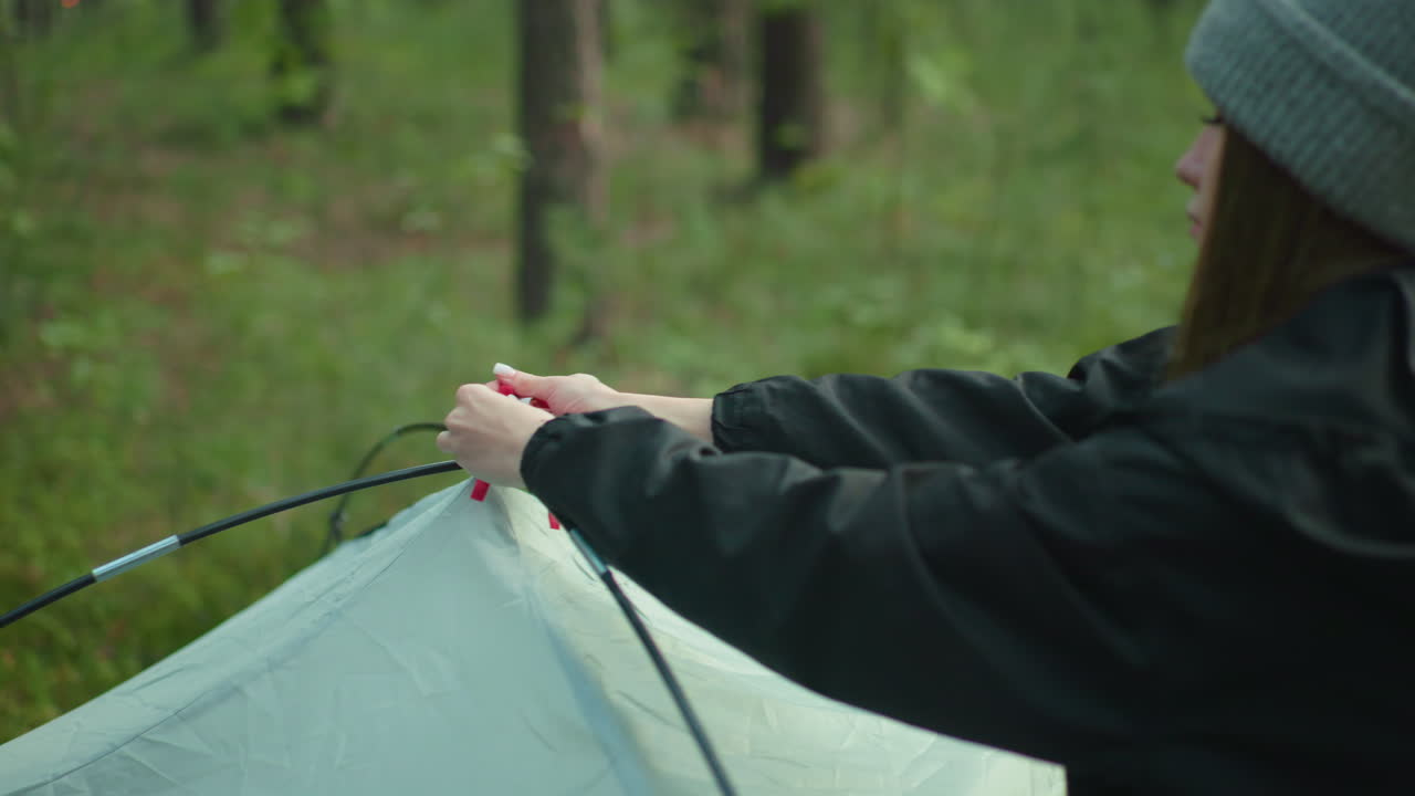 woman in black sweater secures tent fabric to flexible pole during outdoor forest camping setup with tree trunks and green grass creating serene natural background