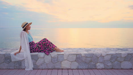 a lone young woman sits on the seawall looking out into the sunset