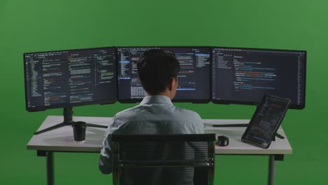 Back View Of Asian Man Developer Working With Tablet While Write Code With Multiple Computer Screens In Green Screen Studio