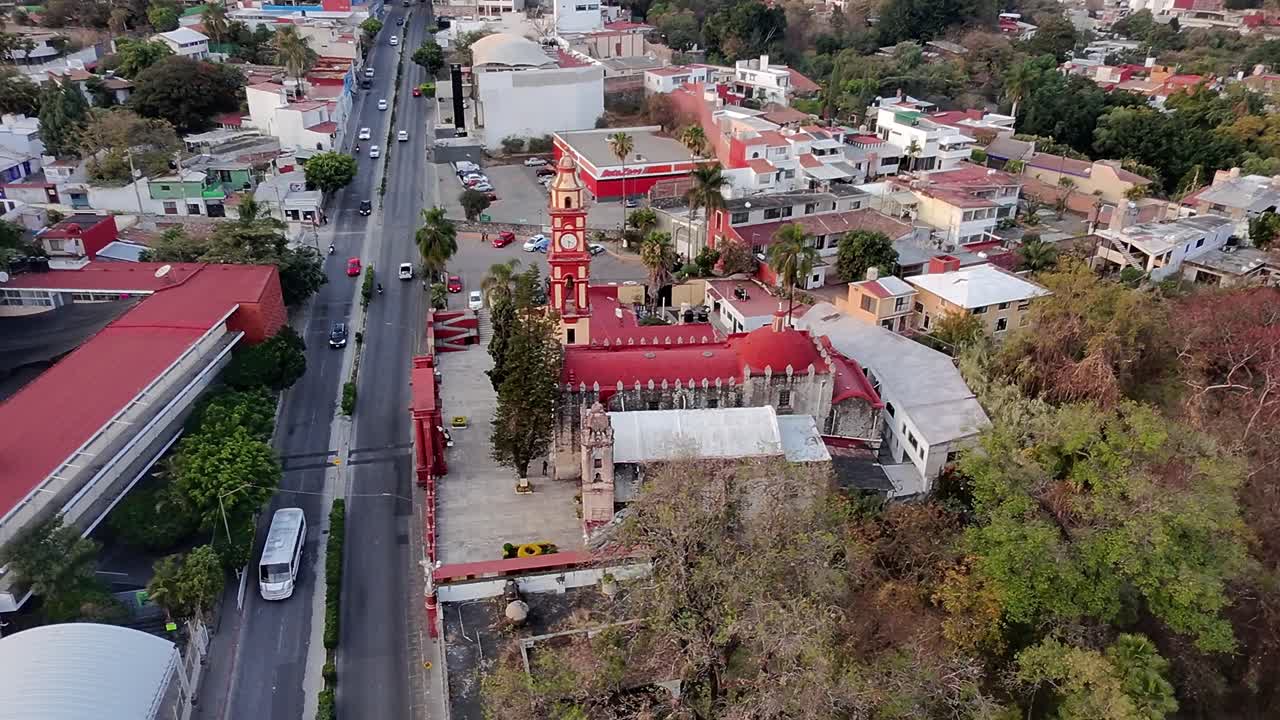 Drone view of Santuario de Tlaltenango with red roof and clock tower, nestled in Cuernavaca’s urban area with nearby homes, busy avenue, and surrounding trees.