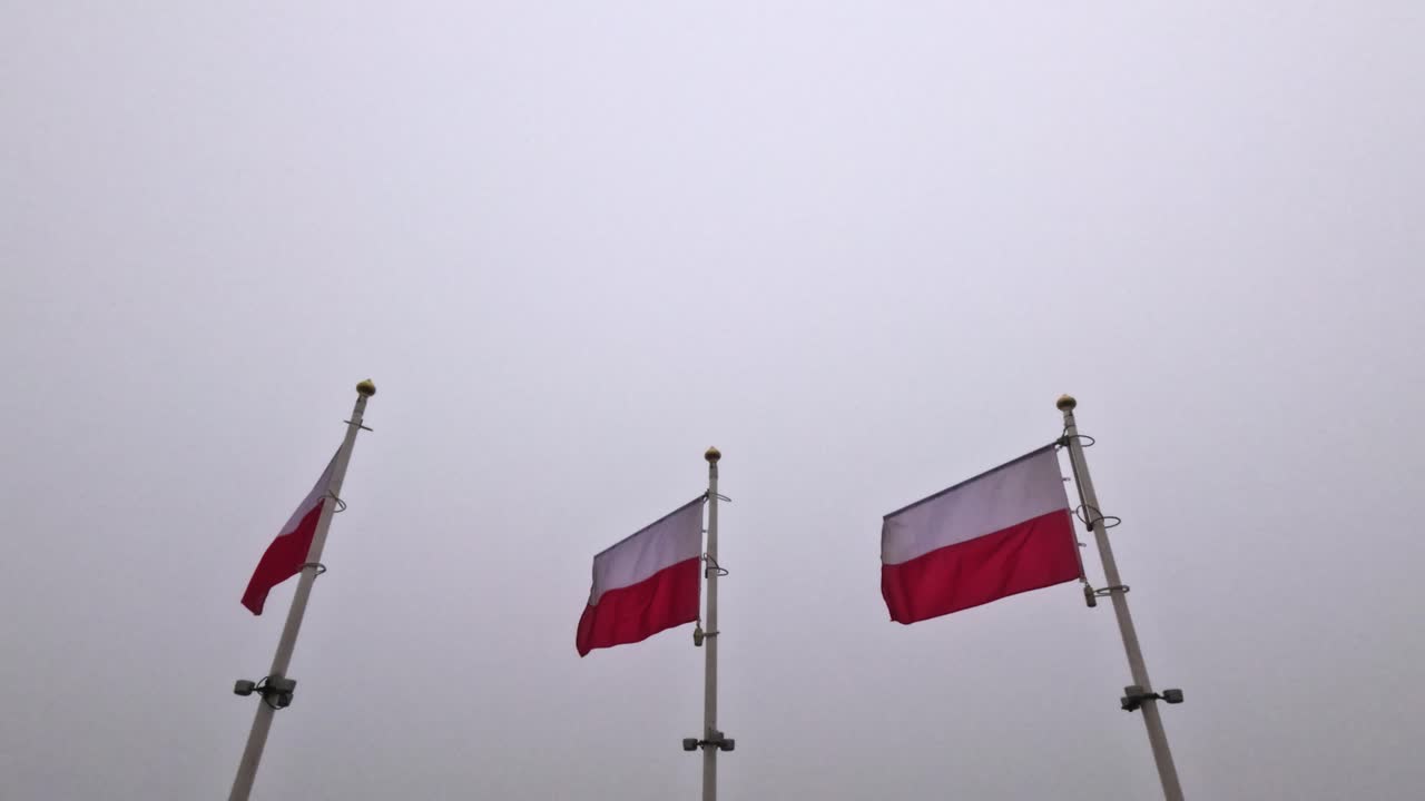 Tiga bendera Polandia dikibarkan di tiang-tiang tinggi di sebuah alun-alun kota. Area tersebut tertutup awan abu-abu, menandakan hari yang berawan. Bendera-bendera tersebut bergerak perlahan tertiup angin.