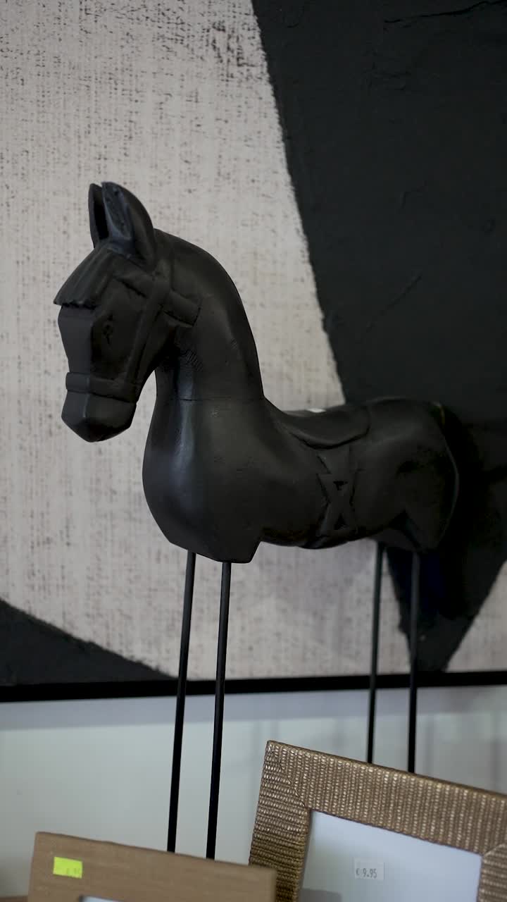 A striking black horse sculpture stands on two metal legs, surrounded by decorative frames. The minimalist art backdrop enhances its elegant design in the room