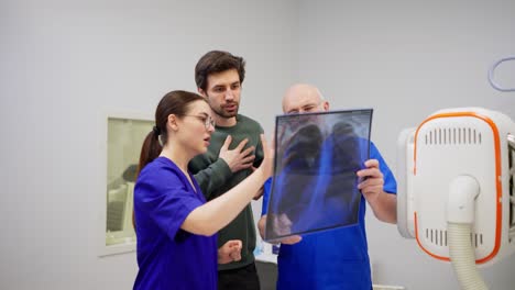 un médico confiado con gafas y barba junto con su asistente una chica morena con un uniforme azul le dice al chico moreno y sostiene una radiografía de pecho delante de ellos en una clínica moderna en la oficina