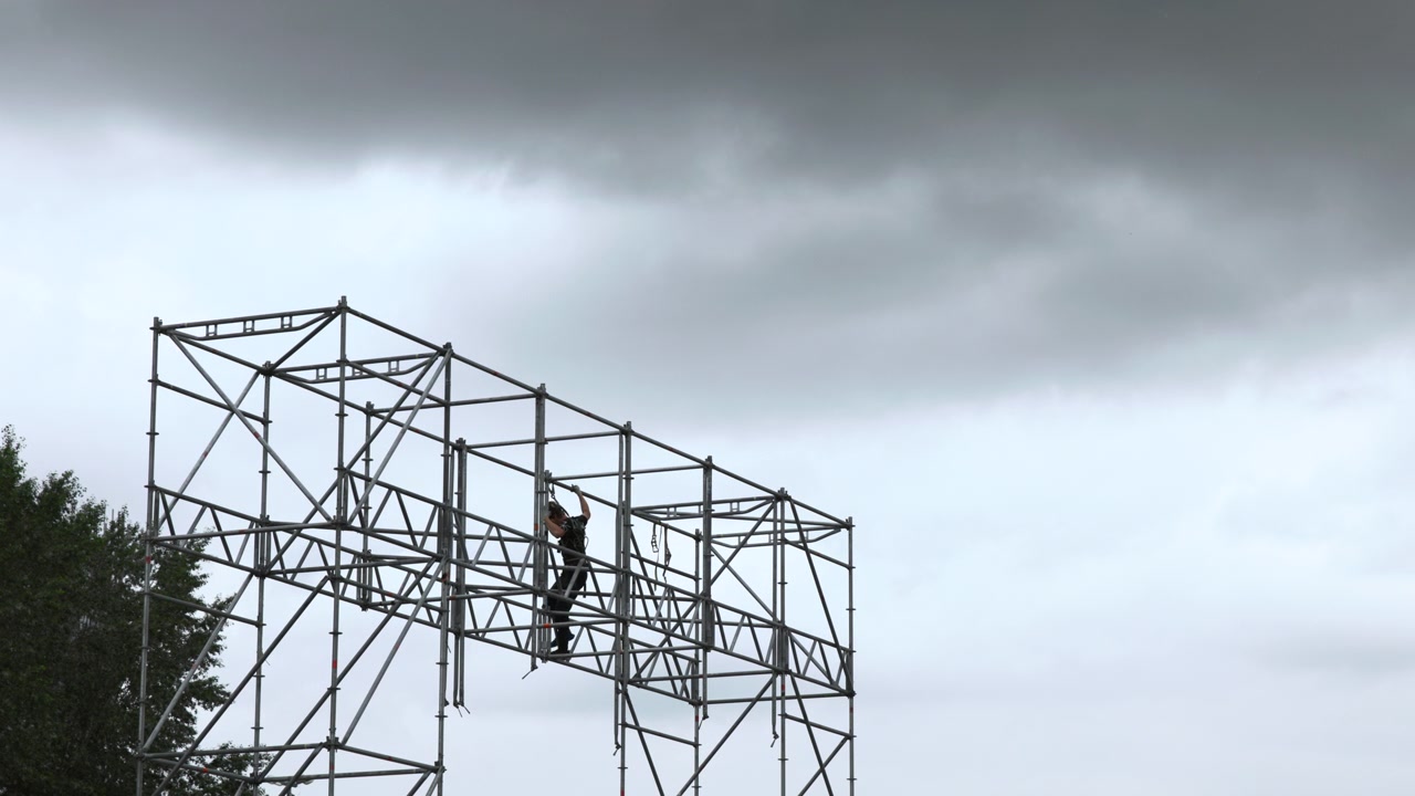 Single construction worker wearing helmet and safety harness stands on modular metal scaffolding, handling ropes and straps while securing bracing under moody overcast sky at elevated industrial site