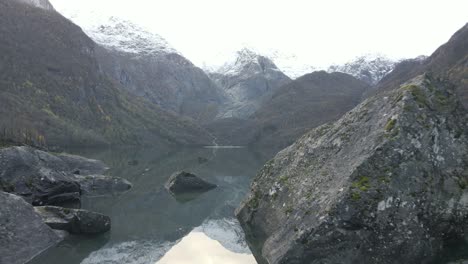 Mountain-lake-with-boulders-and-stunning-mountain-range-in-horizon,-aerial-fly-forward-shot