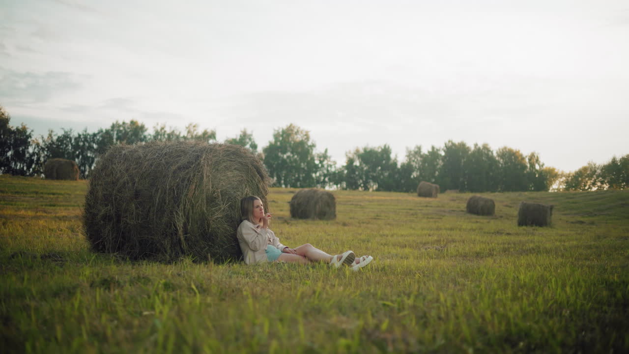 Uma jovem sentada no chão com as pernas cruzadas pensativamente, encostada a um grande fardo de feno em terras agrícolas abertas. Um cenário rural tranquilo com fardos de feno espalhados e luz suave do entardecer.