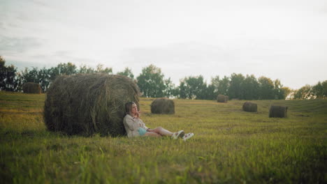 young lady seated on ground with legs crossed thoughtfully, resting back against large hay bale in open farmland, peaceful rural setting with scattered hay bales, soft evening light