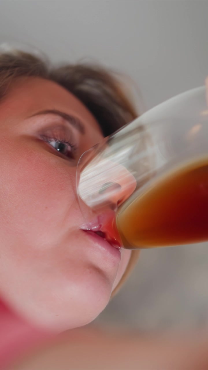 Primo piano di donna che beve da un bicchiere con liquido marrone. La sequenza mostra la progressione delle sue labbra che si muovono verso e lontano dal bordo, catturando sottili espressioni facciali durante il consumo. Luce soffusa.