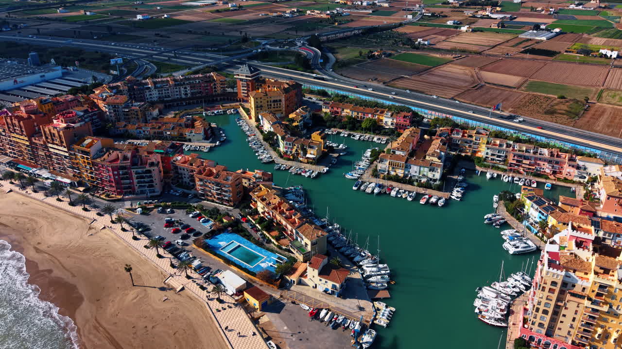 Port Saplaya kystresort med strand og lystbådehavn. Høj vinkel udsigt over farverige lejligheder, sandstrand og både i Port Saplaya marina ved daglys