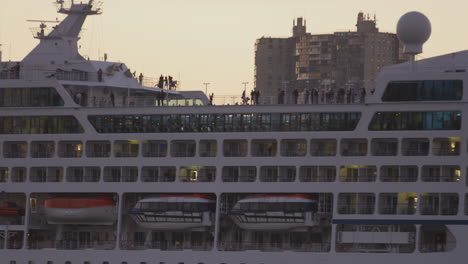 Passengers-Standing-Along-Top-Deck-Of-Cruise-Ship-During-Golden-Hour-With-New-Jersey-Buildings-Seen-In-Background