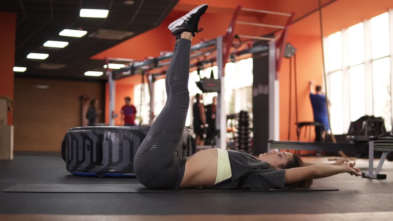 Good looking girl is lifting her torso up to thelegs, lying on floor, tensing abdominal muscles in gymnastic hall. She is keeping fit her body. Side view