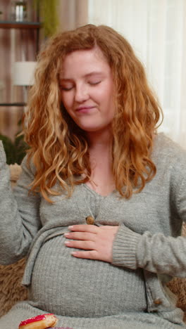 pregnant young woman eating donuts on home sofa laughing with joyful expression enjoying sweet treat