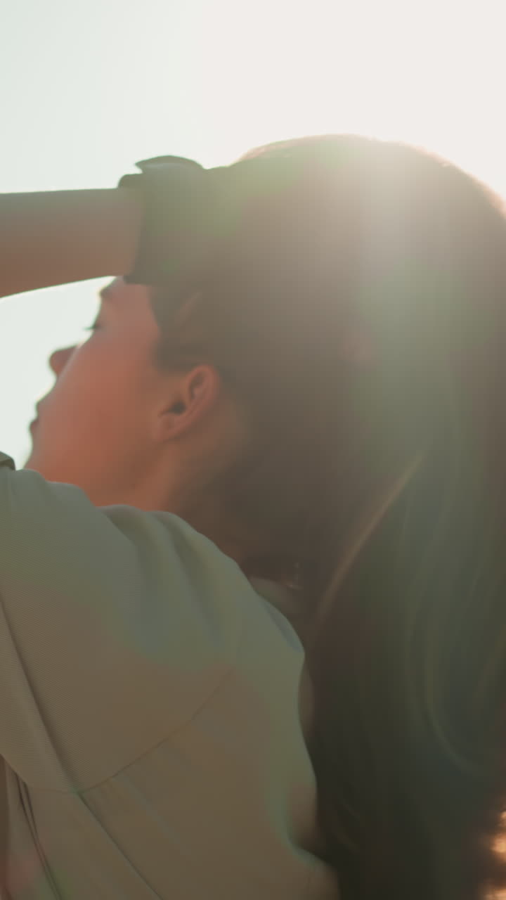 검은 장갑을 입은 여자는 머리를 뒤로 기울이고, 따뜻한 빛 아래 머리를 조정하며, 차분한 초록색 나무와 잎자루가 배경에서 부드럽게 흐릿해집니다.