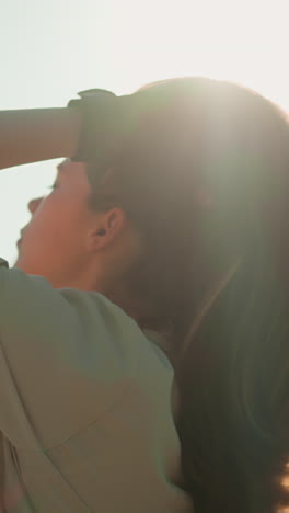 dama con guantes negros inclina la cabeza hacia atrás, ajustando su cabello en la cálida luz del sol, con árboles verdes serenos y follaje suavemente borroso en el fondo