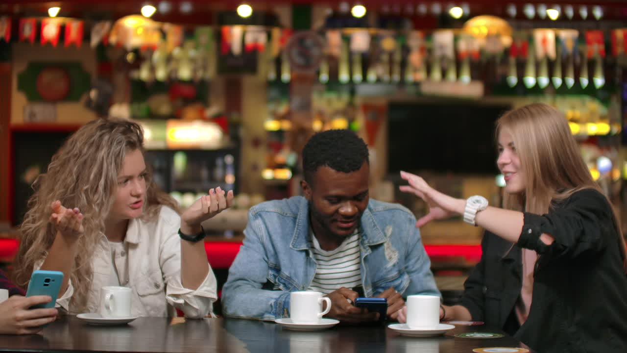 Amici felici e diversi si divertono insieme con un gadget, guardando un video divertente online sullo smartphone; giovani amici multiculturali ridono, guardano il telefono, parlano e usano app mobili seduti al tavolo durante un incontro