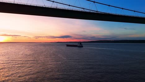 In-this-mesmerizing-drone-clip,-a-barge-boat-drifts-leisurely-beneath-the-Humber-Bridge-at-sunset,-bathed-in-the-golden-hour's-radiance,-while-the-river-sparkles-in-the-setting-sun's-glow