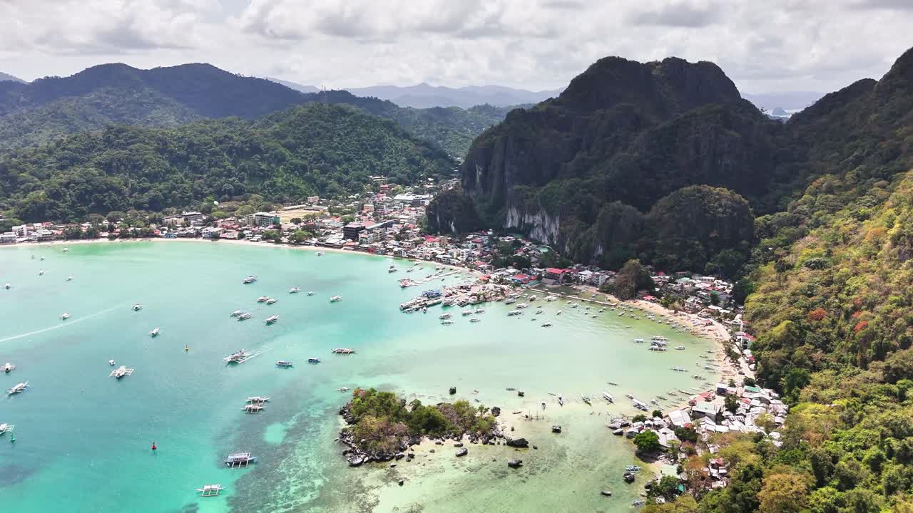 Drone shot of El Nido, Palawan, showing its vibrant coastal town tucked between dramatic karst cliffs and the turquoise waters of Bacuit Bay, surrounded by lush rainforest