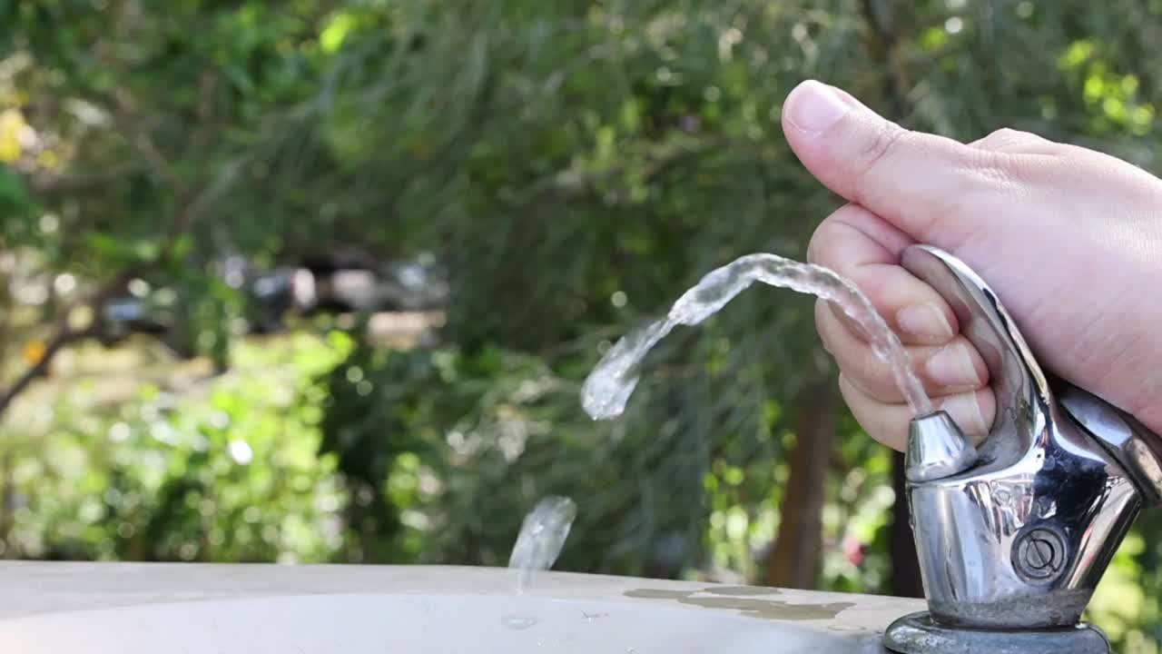 Una mano preme un pulsante di metallo su una fontana da esterno, rilasciando un getto d'acqua