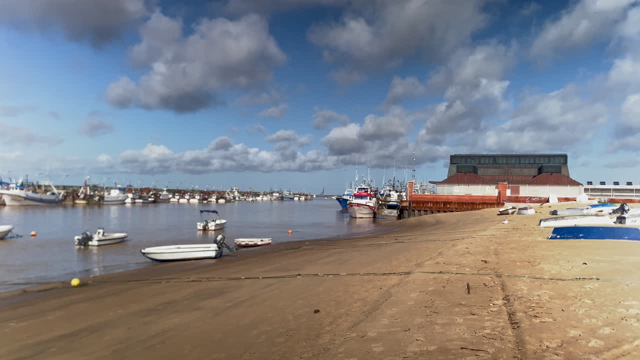 Fishing boats docked in a busy harbor during an overcast day, with small boats resting on the sandy bank