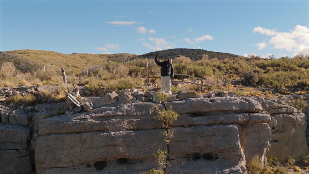 Donna che cerca il segnale del cellulare su una scarpata rocciosa circondata dalle montagne andine, dalla steppa arbustiva e da cieli drammatici, ripresa aerea con orbita del drone, Paso Pehuenche, Malargüe, Mendoza, Argentina.