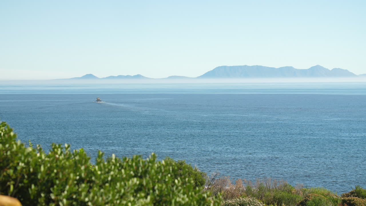 Fishing trawler sailing away in blue waters off the coast of South Africa on clear sunny day with heatwaves and green shrubbery in foreground and misty mountains in background