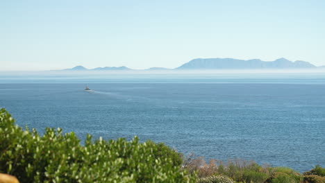 fishing trawler sailing away in blue waters off the coast of south africa on clear sunny day with heatwaves and green shrubbery in foreground and misty mountains in background