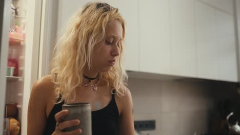 a blonde girl in a black top drinks tea in the morning and looks at her phone in the morning in the kitchen near the refrigerator