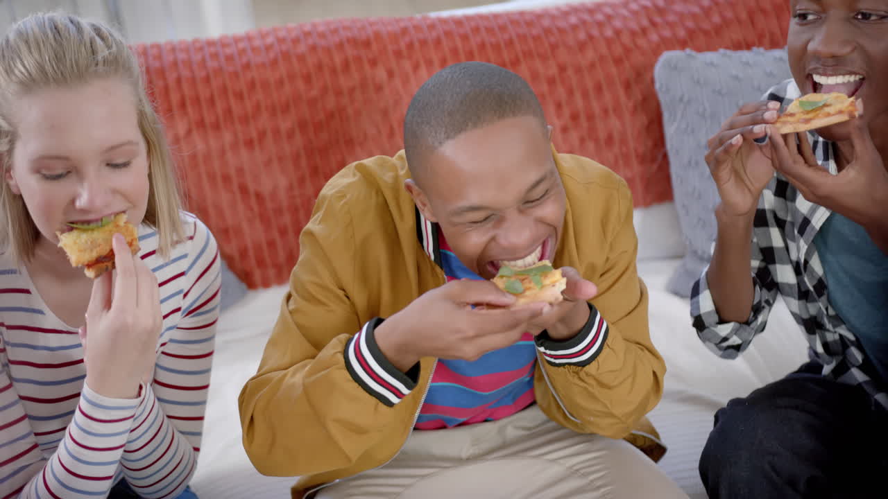 Happy diverse group of teenage friends sitting on couch and eating pizza at home, slow motion