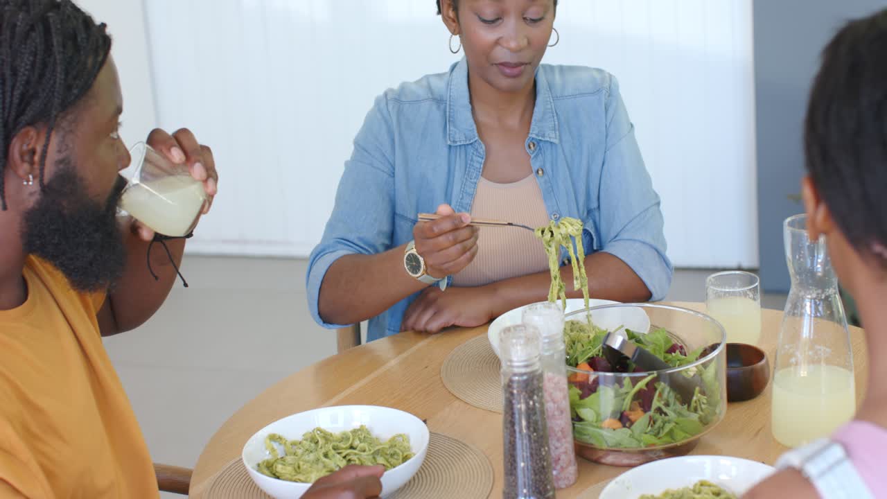 Família afro-americana preparando-se para comer em uma mesa de jantar de madeira, a mulher servindo e enrolando macarrão.