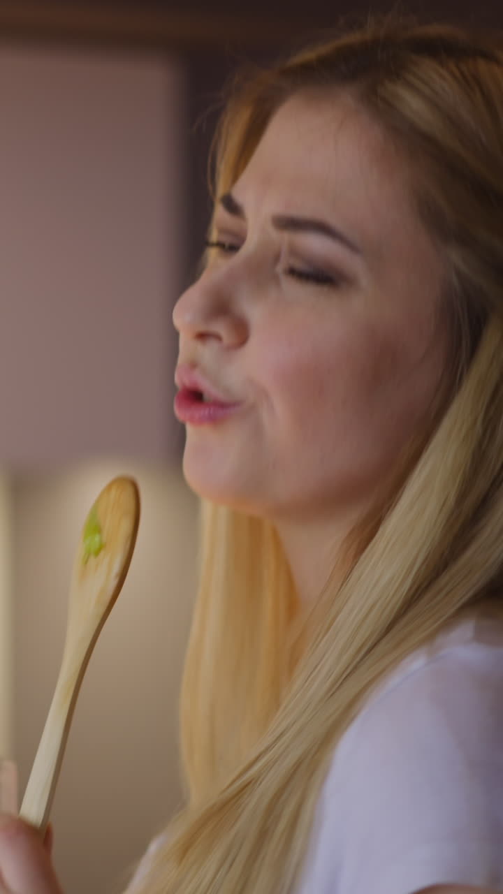 joven rubia positiva en delantal canta una canción y bebe un delicioso vino cocinando una cena festiva en la cocina en casa vista de cerca en cámara lenta