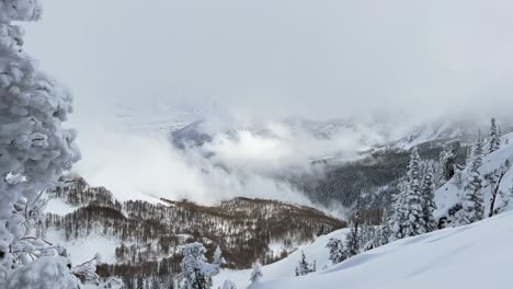 Pan-right-shot-revealing-a-stunning-winter-landscape-scene-looking-down-at-a-cloudy-snow-covered-valley-from-the-summit-of-a-ski-resort-in-the-Rocky-Mountains-of-Utah-on-a-cold-overcast-day