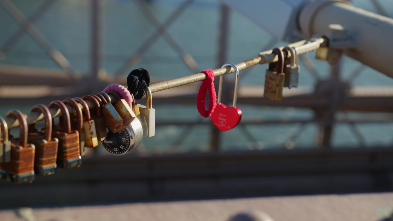 Bright red heart shaped padlock stands out among weathered brass and steel love locks on a bridge railing, the river glittering softly out of focus behind.