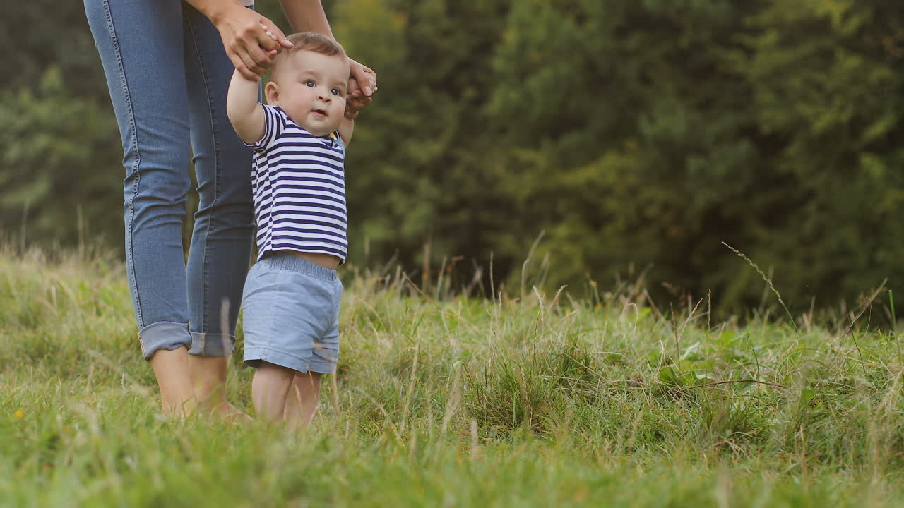 lindo niño pequeño sosteniendo las manos de su madre y dando los primeros pasos descalzos en la hierba