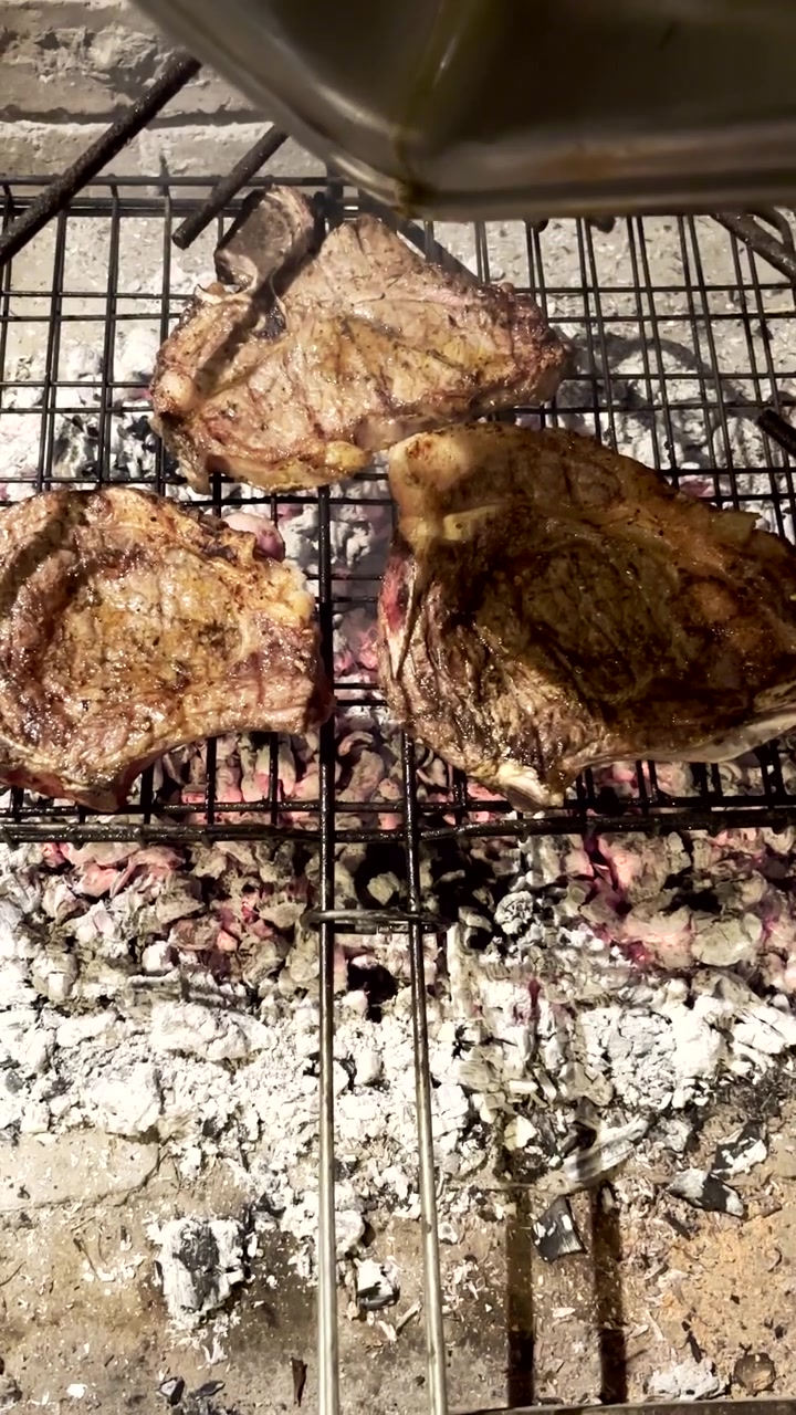Azeite escorrendo sobre bifes T-bone chiando em grelha de malha sobre brasas quentes de madeira. Braai tradicional sul-africano ou churrasco. Vertical.