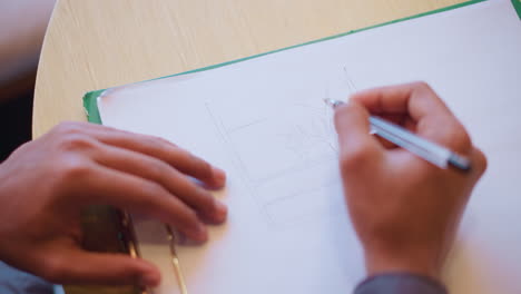person drawing architectural lines with pen on blank white sheet clipped to green folder, using steady hand while seated at table, showing careful technical sketching and concept development phase