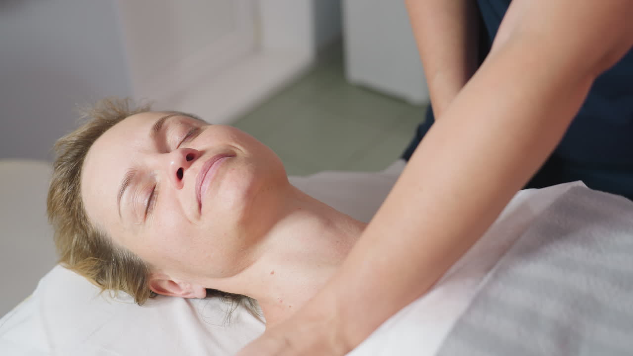 Female client lies on massage bed with eyes closed and serene smile as beauty technician tenderly glides hands along shoulders and neck delivering relaxing wellness session in softly lit spa room