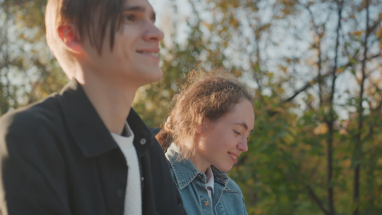 Side profile couple sharing laughter on sunlit park bench, closeup smiles and playful exchange, gentle breeze through trees, candid joyful moment suggesting young family or close partnership