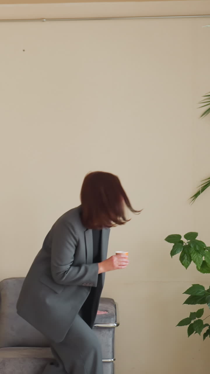 Деловая женщина отходит от дивана в современном офисе, держа в руке напиток, рядом комнатные растения, момент непринужденного перерыва, расслабленная поза, профессиональная одежда, офисная зона отдыха