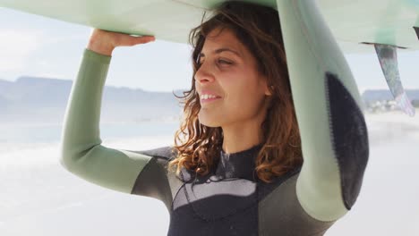 feliz mujer de raza mixta caminando en una playa soleada junto al mar sosteniendo una tabla de surf en la cabeza y sonriendo