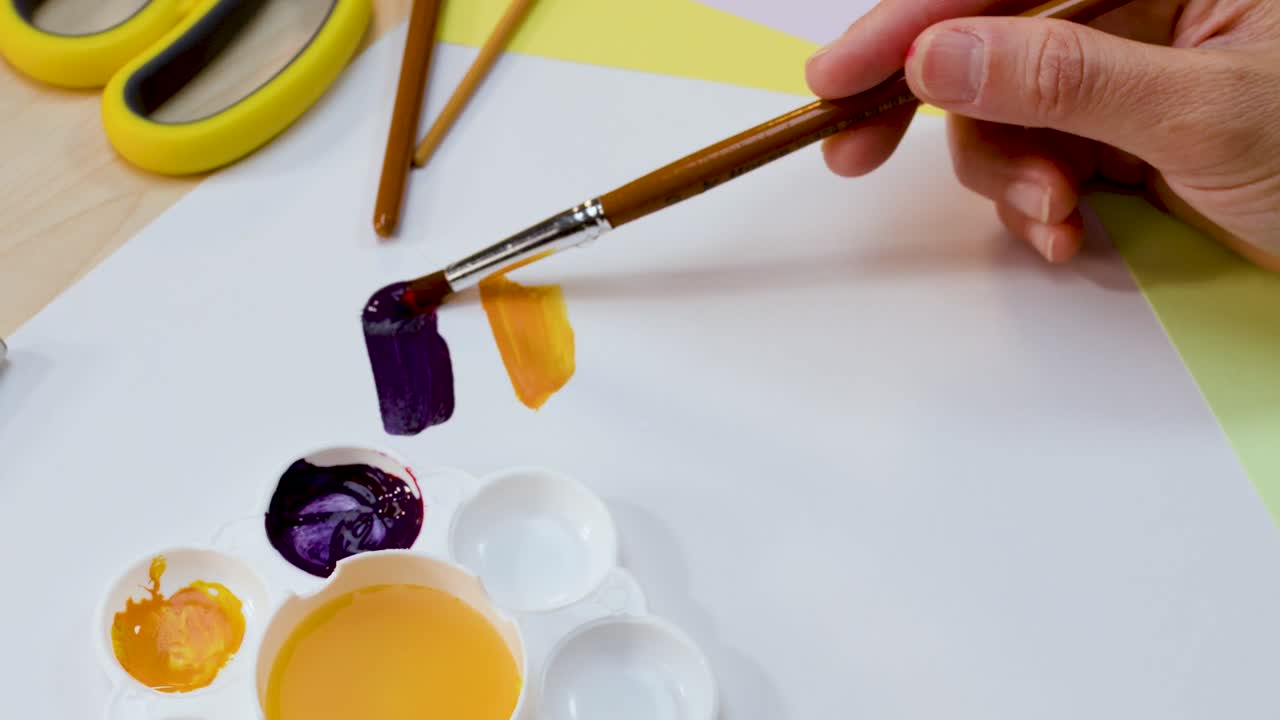 A hand dips a paintbrush into violet watercolor and makes strokes on white paper