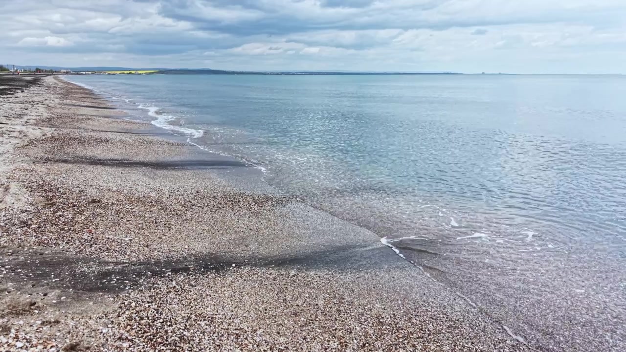Widok na spokojną plażę ze spokojnymi falami i zachmurzonym niebem, ukazujący piasek i wodę pod szarymi chmurami w ciągu dnia