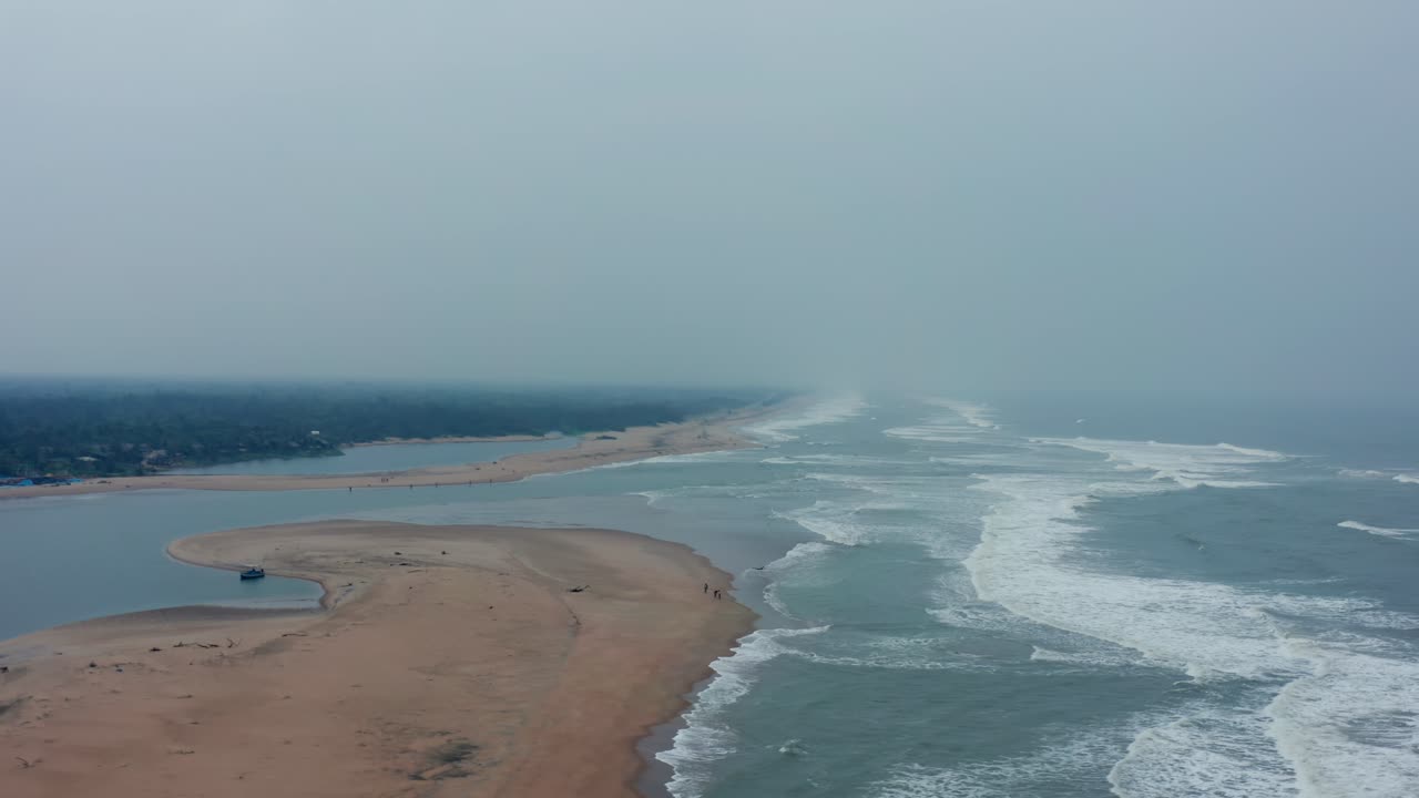 Hazy aerial captures Chandrabhaga Beach's sweeping curved sandbar between river creek and ocean waves, framed by distant green forested banks