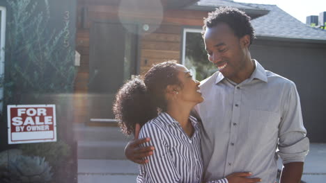 portrait of hugging couple standing outdoors in front of house with for sale sign in garden