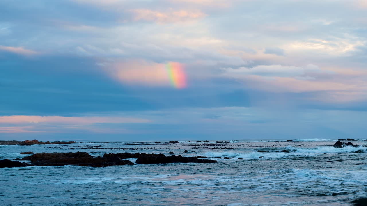 vista sobre las olas y las rocas costeras con arco iris parcial en nubes al atardecer