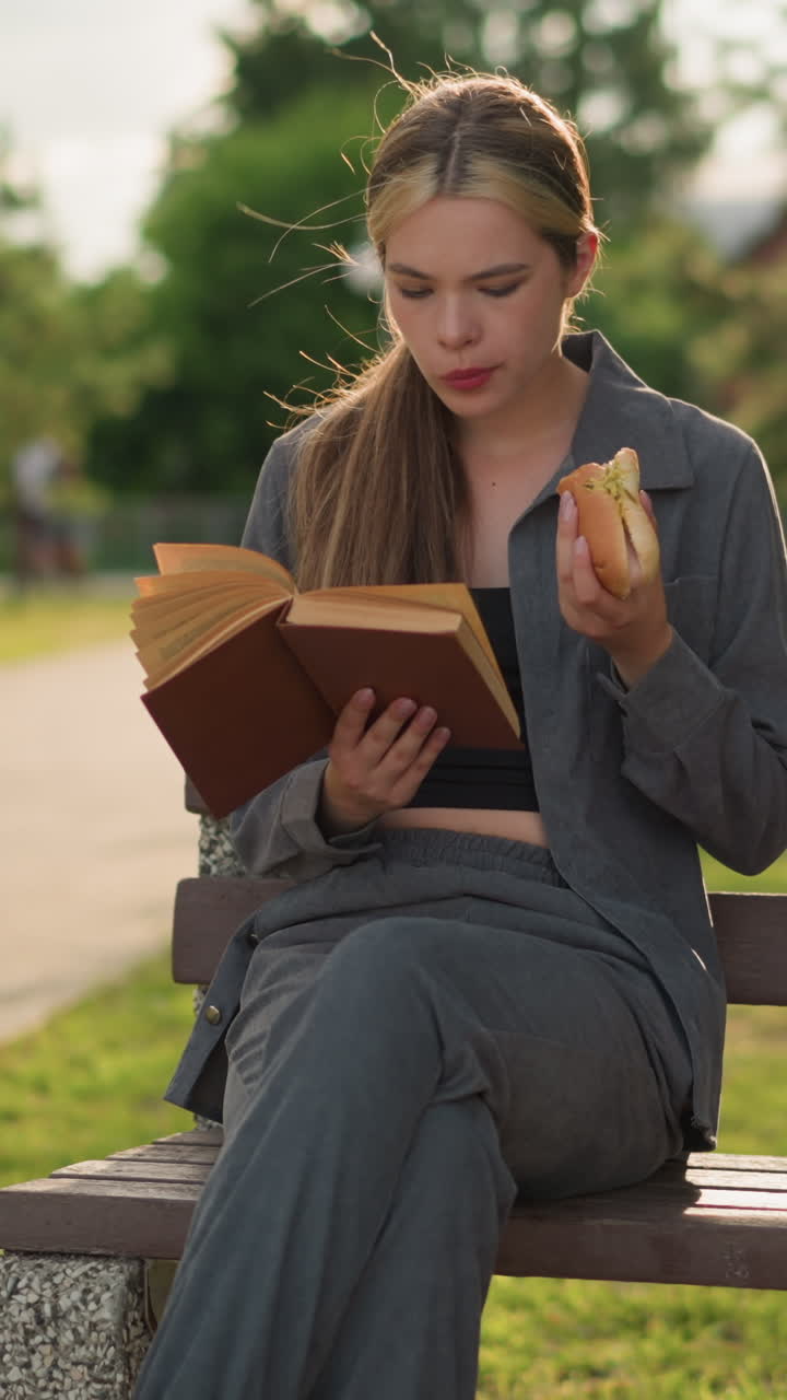 dama comiendo un bocadillo mientras lee un libro al aire libre, sentada en un banco del parque con las piernas cruzadas, disfrutando de un tiempo tranquilo en el parque verde, rodeada de árboles, un camino y un edificio en el fondo