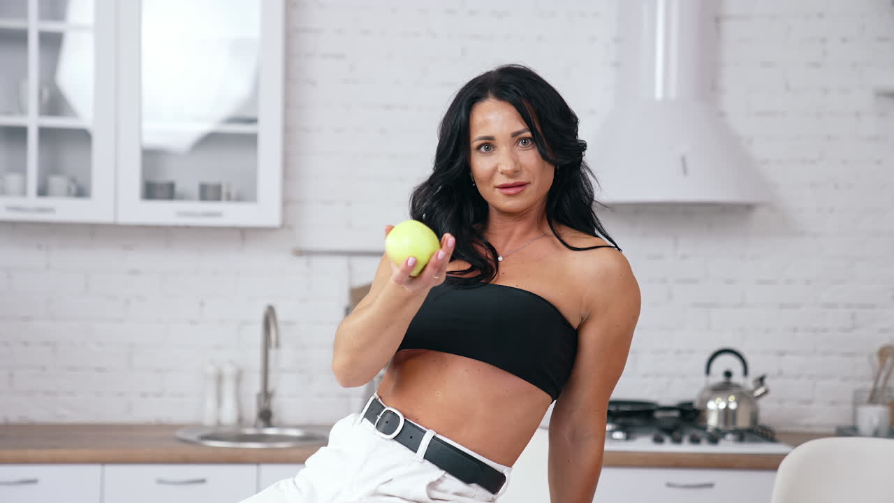 Dark- haired woman with a green apple. Attractive woman sitting on the table in the kitchen and proposing fresh apple. Healthy food.
