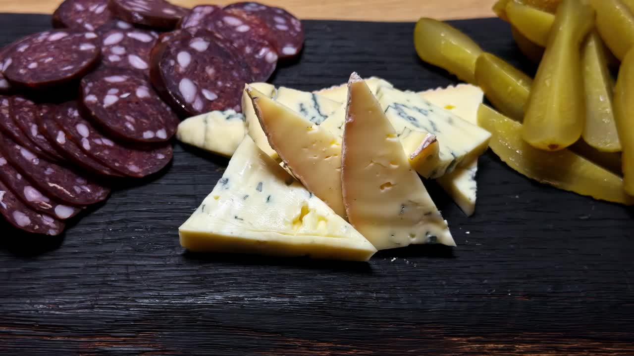 Sliced salami, creamy blue-veined cheese triangles, and crisp pickles arranged on a dark serving board, creating a rustic gourmet snack platter with rich texture and warm, inviting tones